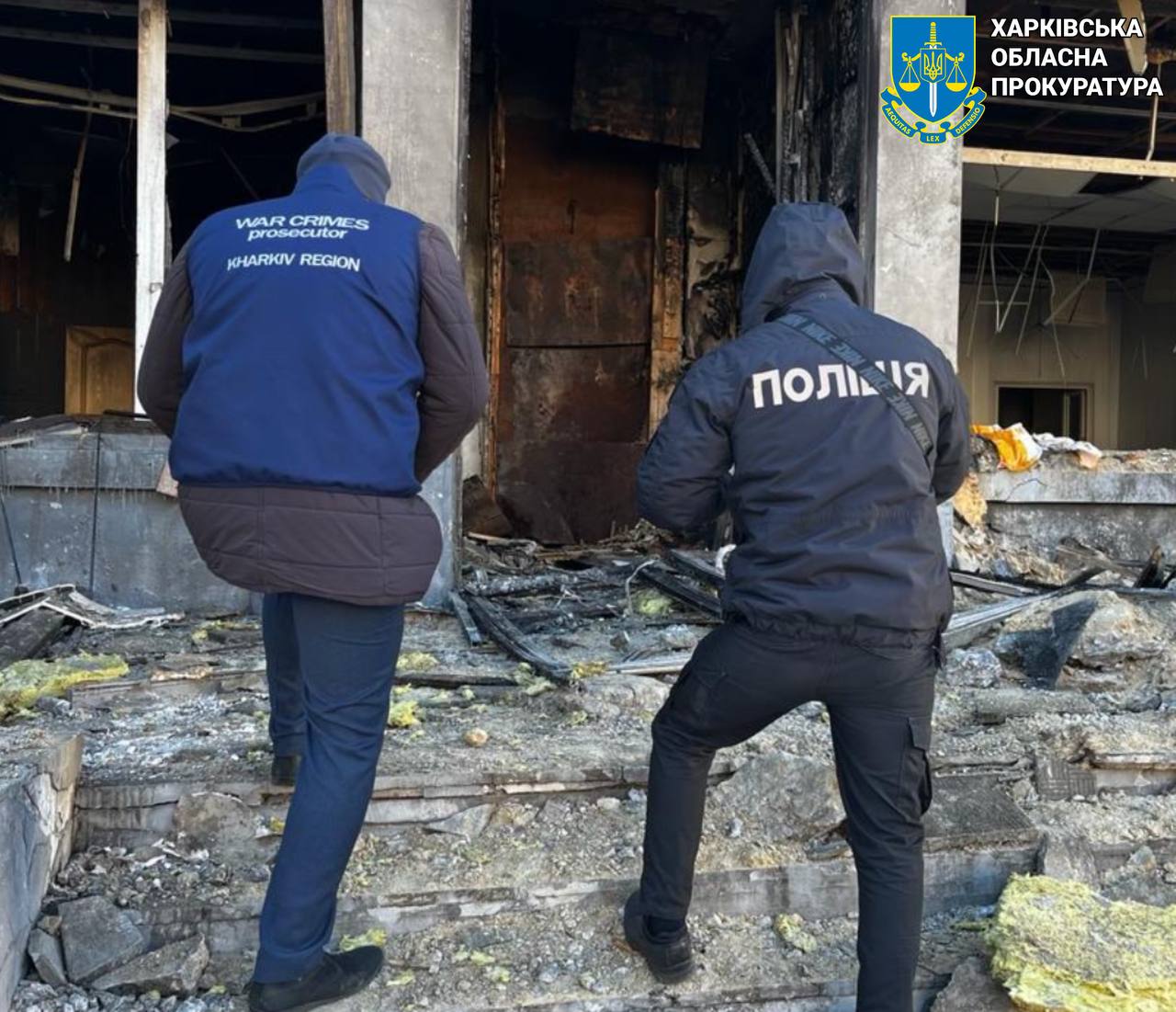 Two men in black jackets standing on stairs outside a burned out building

Description automatically generated