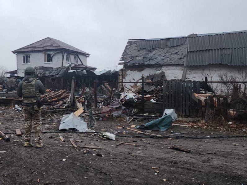 A person in a military uniform standing in front of a destroyed house

Description automatically generated