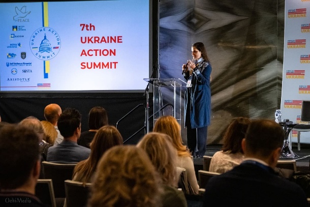 A speaker stands at a podium facing an audience, standing next to a Ukraine Action Summit presentation.