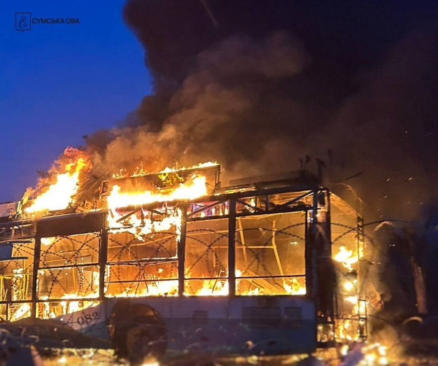 A light-blue and white trolleybus with all its windows shattered is engulfed in flames