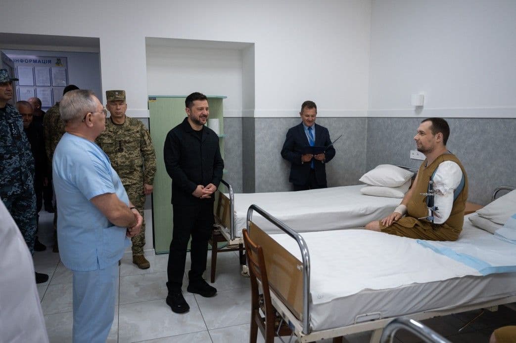 Presisdent Volodymyr Zelensky, wearing all black stands by the bedside of a wounded Ukrainian soldier in a medical facility.